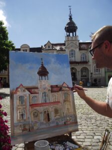 Zdjęcie przedstawia malarza Denisa Chyżawskiego przy pracy w słoneczny dzień. Malarz stoi przy sztaludze, w ręce trzyma pędzel i maluje obraz kościoła, widocznego na drugim planie zdjęcia.