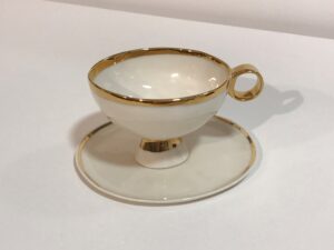 Piękna filiżanka do herbaty z białej porcelany, inspirowana powieścią „Alicja w krainie czarów”, z talerzykiem. Filiżanka jest wykonana z białej porcelany i jest ozdobiona 24-karatowym złotem: złoty pasek u styku czarki z nóżką, wokół rantu filiżanki i krawędzi talerzyka. To wzór klasycznej elegancji. Produkt dostępny w Galerii Wiele Sztuki.