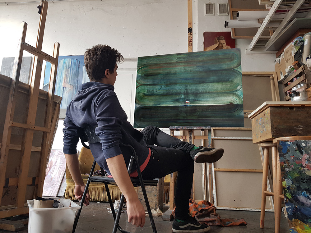 Zdjęcie profilowe artysty malarza Allena Macka do biogramu na stronie internetowej Galerii Wiele Sztuki. Malarz siedzi na krześle w swojej pracowni przed ukończonym obrazem. 