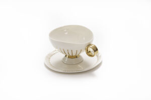 Piękna filiżanka do herbaty z białej porcelany, inspirowana powieścią „Alicja w krainie czarów”. Filiżanka jest wykonana z białej porcelany i jest niezwykle ciekawie zdobiona złoceniami z 24-karatowego złota: ma bogato pokryte złotem uszko, na korpusie ma złote paseczki zaczynające się małą złotą kroplą biegnące w dół do górnej podstawy nóżki. Na górnej podstawie nóżki jest gruby pasek złota. W komplecie do filiżanki jest talerzyk z białej, szkliwionej porcelany ozdobiony małymi paseczkami, biegnącymi od brzegu talerzyka w kierunku środka. Produkt dostępny w Galerii Wiele Sztuki.