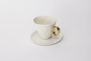 Piękna, biała filiżanka o pojemności 220 ml, z subtelnymi zdobieniami z 24-karatowego złota w formie złotych kropli umieszczonych na korpusie i pozłacanym uchwytem, z serii Piegowata Księżniczka. W komplecie z filiżanką jest talerzyk z białej, szkliwionej porcelany. Produkt dostępny w Galerii Wiele Sztuki.