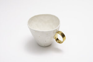 Piękny kubek z białej porcelany, z subtelnymi złoceniami z 24-karatowego złota w postaci małych złotych kropli na korpusie i złotym uszkiem. Produkt dostępny w Galerii Wiele Sztuki.