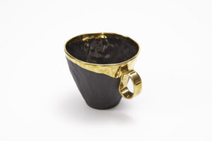 Piękny kubek z czarnej, matowo szkliwionej hiszpańskiej porcelany, z 24-karatowym złotem okalającym rant kubka, a następnie płynnie przechodzącym na uszko. Produkt dostępny w Galerii Wiele Sztuki.
