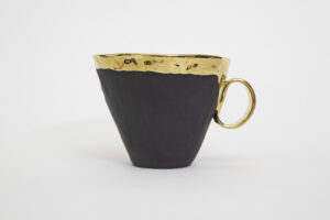 Piękny, elegancki kubek z czarnej, matowo szkliwionej hiszpańskiej porcelany, z 24-karatowym złotem okalającym rant kubka, a następnie płynnie przechodzącym na uszko. Widok na kubek z boku. Produkt dostępny w Galerii Wiele Sztuki.