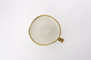 Piękny kubek z białej porcelany, z wyraźnym złoceniem na brzegu i złotym uszkiem. Widok na kubek od góry. Produkt dostępny w Galerii Wiele Sztuki.