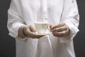 Kobieta trzyma w rękach piękną, białą, złoconą filiżankę z talerzykiem, Filiżanka jest wykonana z białej porcelany AUDREY BLACKMAN znanej z gładkości, pięknej bieli i przejrzystości. Filiżanka jest dostępna w Galerii Wiele Sztuki.