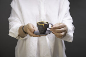 Kobieta trzyma w rękach piękną, czarną, złoconą filiżankę z talerzykiem, Zdobienie tej unikatowej, czarnej porcelany to 24-karatowe złoto, które otoczyło cały rant, a spadając delikatną kroplą na pozostawiło ślad na boku filiżanki. Produkt dostępny w Galerii Wiele Sztuki.