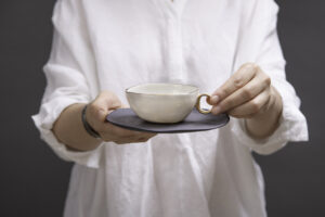 Kobieta trzyma w rękach piękną, filigranową filiżankę do herbaty z białej porcelany z delikatnie złoconym rantem i złoconym uszkiem. W komplecie z filiżanką jest talerzyk z czarnej matowej porcelany z subtelnymi złotymi zdobieniami w formie małych kropli. Złocenia wykonane z 24-karatowego złota. Produkt dostępny w Galerii Wiele Sztuki.