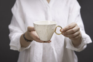 Kobieta ubrana na biało trzyma w rękach piękny kubek z białej porcelany, z subtelnymi złoceniami z 24-karatowego złota w postaci małych złotych kropli na korpusie i złotym uszkiem. Produkt dostępny w Galerii Wiele Sztuki.