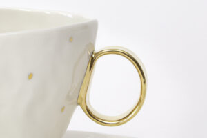 Zbliżenie fragmentu pięknej, białej filiżanki o pojemności 220 ml, z subtelnymi zdobieniami z 24-karatowego złota w formie złotych kropli umieszczonych na korpusie i pozłacanym uchwytem, z serii Piegowata Księżniczka. W komplecie z filiżanką jest talerzyk z białej, szkliwionej porcelany. Na zbliżeniu widać dobrze bogato pokryte złotem ucho filiżanki i subtelne, złote krople na jej korpusie. Produkt dostępny w Galerii Wiele Sztuki.