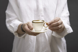 Kobieta trzyma w rękach piękną, białą, złoconą filiżankę z talerzykiem, Filiżanka jest wykonana z białej porcelany AUDREY BLACKMAN znanej z gładkości, pięknej bieli i przejrzystości. Filiżanka jest dostępna w Galerii Wiele Sztuki.