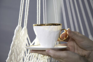 Kobieta trzyma w rękach piękną, filigranową filiżankę do herbaty z białej porcelany ze złoceniami. W komplecie z filiżanką jest talerzyk z białej porcelany ze zdobieniem w formie złotej opaski wokół krawędzi talerzyka. Produkt dostępny w Galerii Wiele Sztuki.