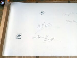 Na zdjęciu jest widoczne odwrocie (tylna strona) rysunku autorstwa Anny Malinowskiej pod tytułem „Fruwające arlekiny”. Widać odręczny podpis artystki, imię i nazwisko, tytuł pracy po polsku i angielsku, rok powstania oraz dwie prostokątne pieczęcie autorskie. Praca jest dostępna w Galerii Wiele Sztuki.