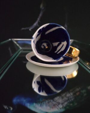 Piękna, biało-kobaltowa filiżanka do espresso o pojemności 60 ml z serii Moja własne galaktyka, ozdobiona 24-karatowym złotem w postaci złoconego uszka i małych subtelnych kropek wewnątrz filiżanki. Filiżanka leży na boku na talerzyku tak, że widać kobaltowe zdobienia wnętrza naczynia i złotą kropkę na jej dnie. Produkt jest dostępny w Galerii Wiele Sztuki.