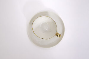 Piękna, biała filiżanka o pojemności 200 ml, z subtelnymi zdobieniami z 24-karatowego złota w formie delikatnej linii wokół krawędzi i złoconym uszkiem. W komplecie z filiżanką jest talerzyk z białej, szkliwionej porcelany. Widok na filiżankę i talerzyk od góry. Filiżanka jest dostępna w Galerii Wiele Sztuki.