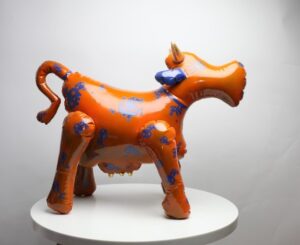 Kolekcjonerska rzeźba ceramiczna 'Krówka mumu - model woodoo' Mariusza Dydo, pomarańczowa figura krowy z niebieskimi motywami czaszek i złotymi rogami, Galeria Wiele Sztuki.